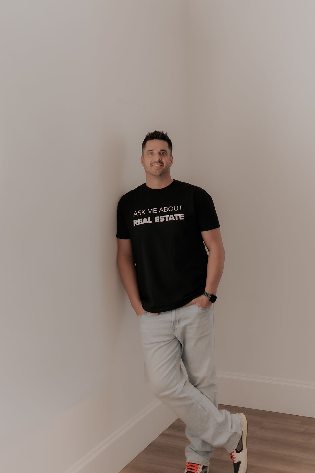 Taylor M Smith leaning against a wall in an Ask Me About Real Estate shirt
